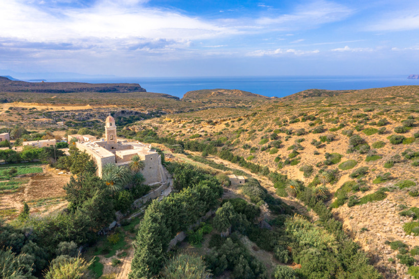 Historisches Kloster mit Glockenturm inmitten einer trockenen, hügeligen Landschaft mit spärlicher Vegetation. Im Hintergrund ist das Meer unter leicht bewölktem Himmel sichtbar