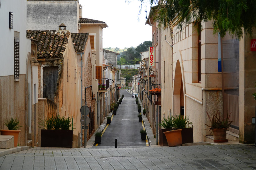 Schmale Altstadtstraße auf Mallorca mit traditionellen Natursteinhäusern, Fensterläden, Pflanzkübeln und Blick auf eine ruhige Dorfstraße