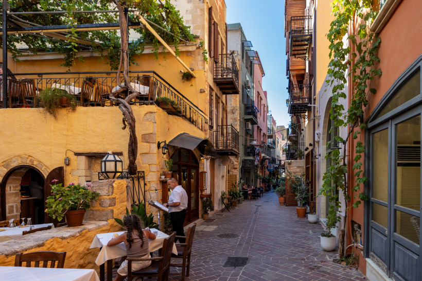 Gasse in der Altstadt von Chania mit Cafés und Balkonen Schmale Gasse in der Altstadt von Chania mit Cafétischen, Balkonen und Kletterpflanzen an den Fassaden