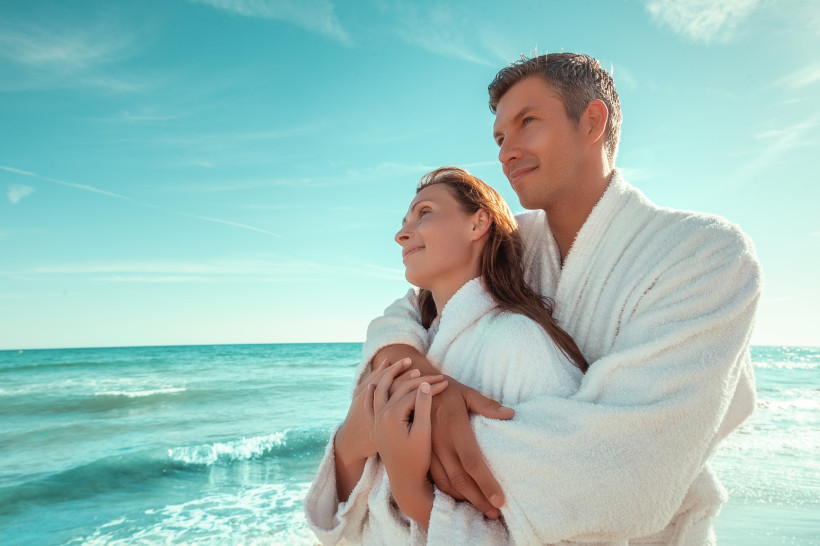 Paar in weißen Bademänteln entspannt am Strand und blickt aufs Mee