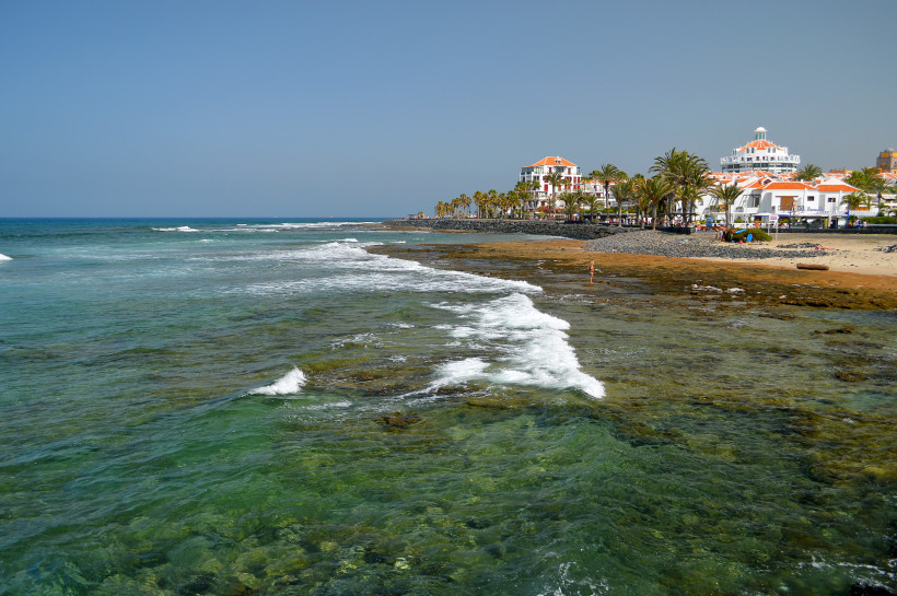 Küstenabschnitt bei Arona auf Teneriffa mit flachem Felsriff, Brandung, Palmen und Gebäuden am Ufer
