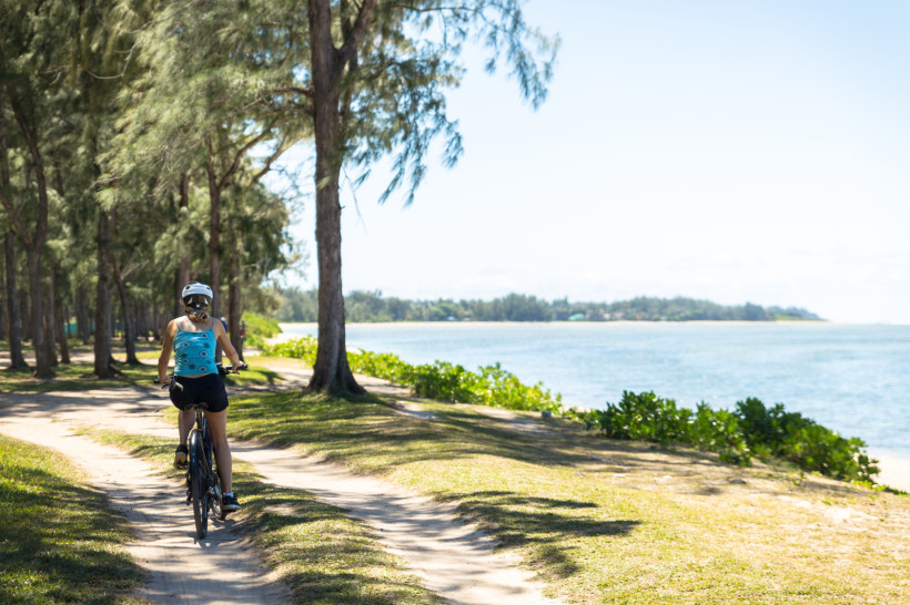 Dieses Bild zeigt eine entspannte Fahrradtour entlang eines malerischen Küstenpfades. Eine Frau mit Helm fährt auf einem schmalen Sandweg, der sich zwischen hohen, schattenspendenden Bäumen und der ruhigen, blauen Küste erstreckt. Das Sonnenlicht fällt sa