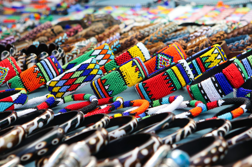 Südafrika Nahaufnahme eines Marktstandes mit vielen farbenfrohen, handgefertigten Armbändern aus Perlen in traditionellen Mustern und Farben. Im Vordergrund sind auch Schnitzarmbänder zu sehen.