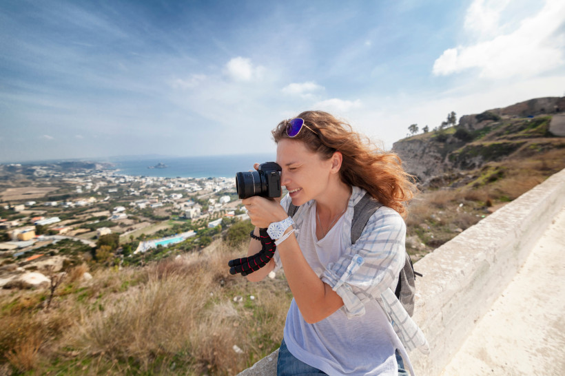 Kos Junge Frau mit Kamera am Auge fotografiert eine weite Landschaft auf Kos. Im Hintergrund liegt eine weite Küstenlandschaft mit Häusern, Feldern und dem Meer. Die Frau trägt Freizeitkleidung, hat windzerzaustes Haar und wirkt konzentriert und glücklich.