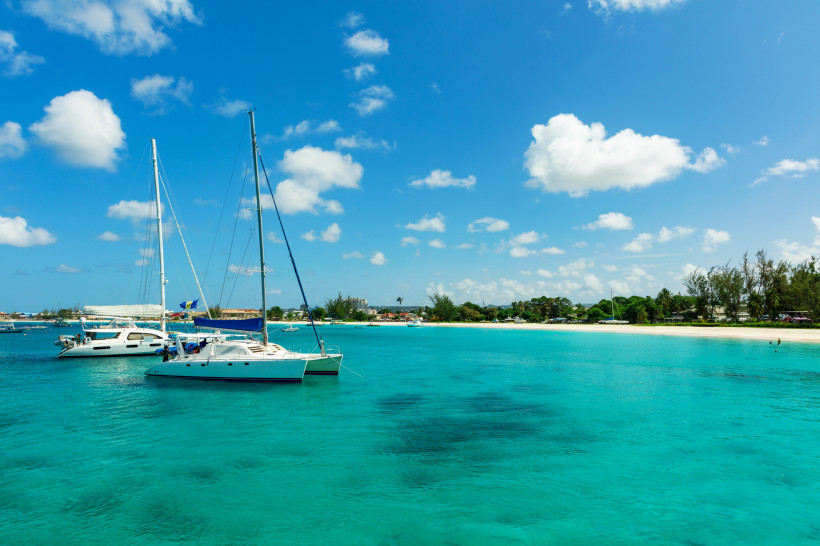 Barbados Zwei weiße Katamarane liegen vor Anker im türkisblauen Meer vor der Küste von Barbados. Im Hintergrund ist der Strand mit Palmen, Häusern und kleinen Booten zu sehen. Der Himmel ist strahlend blau mit wenigen weißen Wolken – perfekte Bedingungen für einen