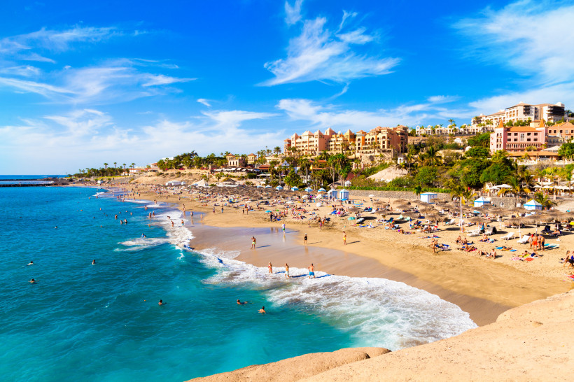 Playa del Duque – exklusiver Strand an der Costa Adeje Blick auf den feinsandigen Strand Playa del Duque an der Costa Adeje auf Teneriffa. Urlauber baden im türkisblauen Meer und sonnen sich am Strand, im Hintergrund luxuriöse Hotels und Palmen.