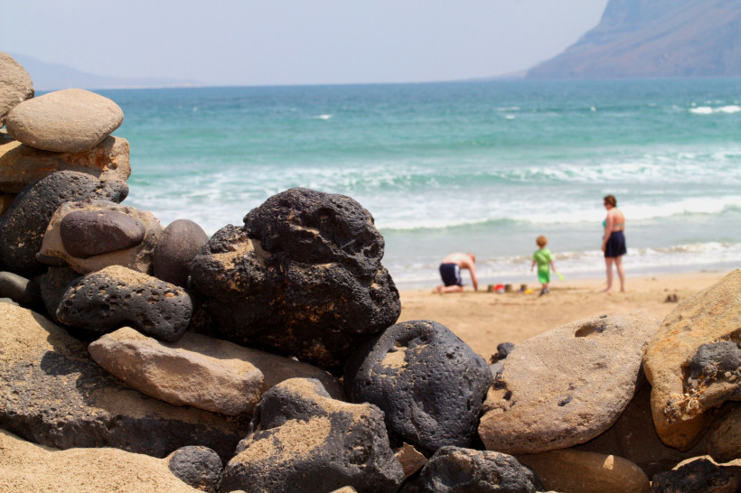 Familie am Strand Lavagestein im Vordergrund an einem Strand, im Hintergrund Familie mit Kind beim Spielen am Meer mit sanften Wellen und Bergküste.