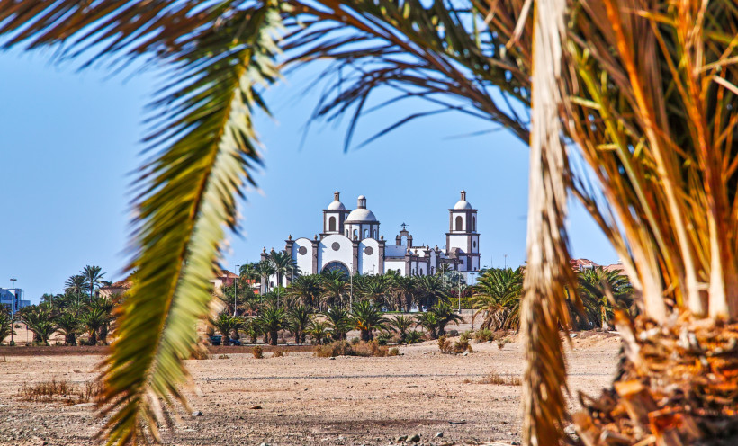 Blick auf eine weiße Kirche mit Kuppeln und Türmen in Meloneras, eingerahmt von Palmen