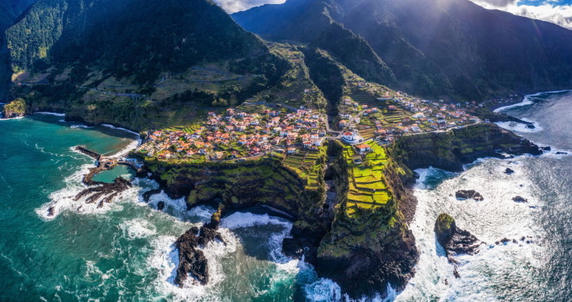 Madeira Das Foto zeigt Felsterrassen mit kleinen Häusern und Feldern, die steil bis zum Meer abfallen.      Zwischen Lavafelsen bricht sich die kräftige Brandung; Berge bilden die dunkle, wolkenumspielte Kulisse.