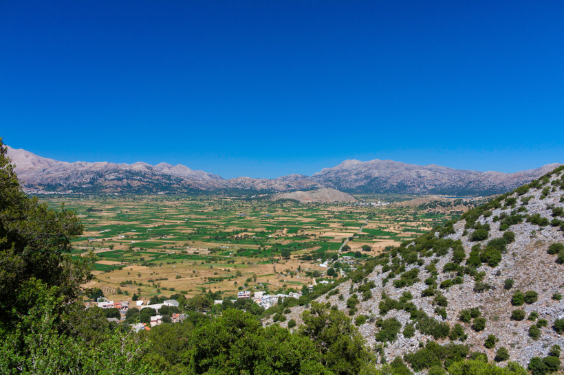Blick über die Lasithi-Hochebene mit Feldern und Dörfern vor einem Gebirgszug unter blauem Himmel
