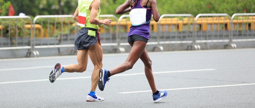 Zwei Marathonläufer:innen in Sportkleidung laufen nebeneinander auf einer abgesperrten Straße. Der Fokus liegt auf ihren Beinen in Bewegung, im Hintergrund sind Absperrgitter und Grünanlagen zu sehen.