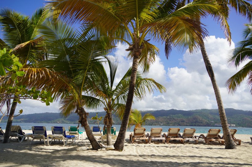 Cayo Levantado, Dom.Rep. Sonnenliegen im Schatten von Palmen am Strand von Samaná in der Dominikanischen Republik mit Blick auf das Meer und die grünen Hügel im Hintergrund