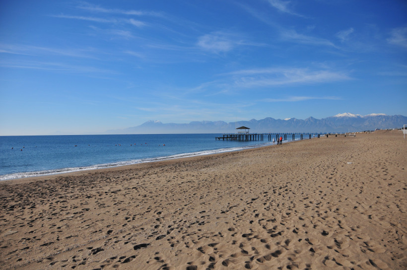 Strand von Kundu an der Türkischen Riviera – Weitläufiger Sandstrand mit Blick auf das Taurusgebirge Weitläufiger Sandstrand in Kundu an der Türkischen Riviera mit ruhigem Meer, Badesteg und Blick auf das Taurusgebirge bei klarem Himmel.