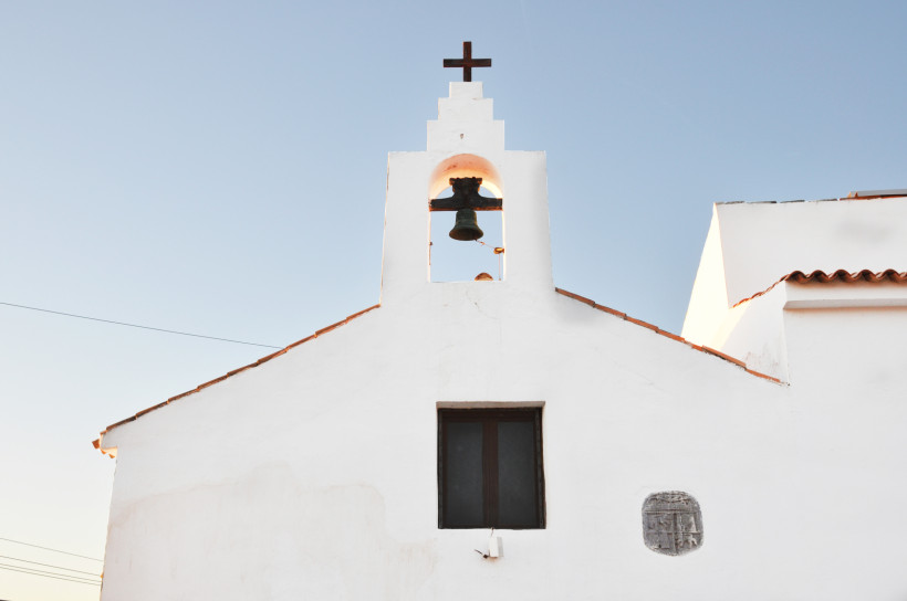 Ibiza Eine kleine weiße Kirche mit einfacher Fassade, einem dunklen Fenster und einem Glockengiebel mit Kreuz auf dem Dach. Die Glocke hängt frei sichtbar unter dem blauen Himmel. Die Architektur wirkt minimalistisch und traditionell, typisch für den Mittelmeer