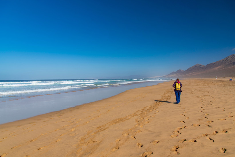 Sandstrand der Playa de Cofete mit Brandung, blauem Himmel und einer wandernden Person am Ufer