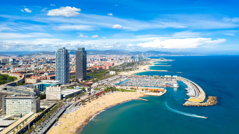 Luftaufnahme von Barcelona mit Blick auf die Küste, den langen Sandstrand, die Marina Port Olímpic und die markanten Hochhäuser Torre Mapfre und Hotel Arts. Im Hintergrund die Stadt und umliegende Berge unter blauem Himmel.