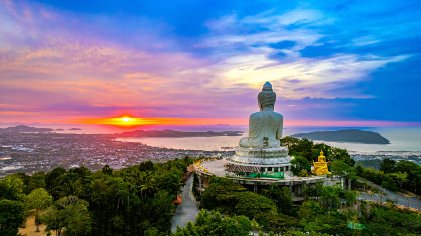Thailand - Phuket Panoramablick auf die berühmte weiße Buddha-Statue auf dem Nakkerd Hill in Phuket, Thailand. Die Statue blickt auf die Stadt und das Meer hinaus, während im Hintergrund die Sonne spektakulär untergeht. Der Himmel ist in kräftigen Farben gefärbt – von Oran