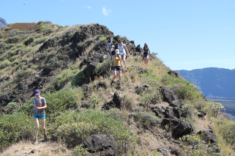 Eine Gruppe von Wandernden steigt einen schmalen, felsigen Pfad in einer hügeligen, trockenen Landschaft mit niedriger Vegetation hinab. Die Szene wirkt sonnig und offen, im Hintergrund sind steile Felswände zu sehen.