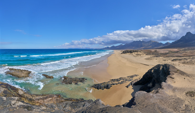 Endloser Naturstrand mit rauer Brandung, Playa de Cofete – Fuerteventura Langer, leerer Naturstrand mit Wellen, türkisfarbenem Meer, Lavafelsen, Dünenlandschaft und Vulkanbergen im Hintergrund unter blauem Himmel