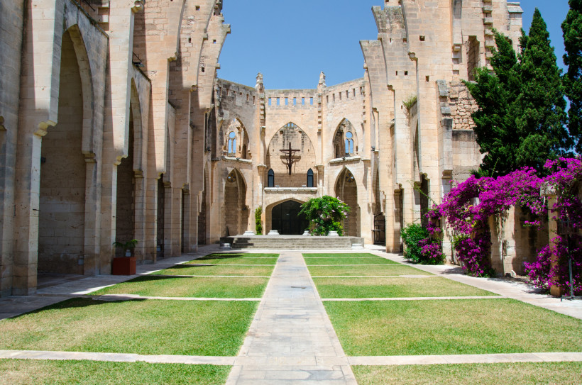 Innenhof einer gotischen Kirche auf Mallorca mit steinernen Arkaden, Kreuz im Altarbereich, symmetrischen Rasenflächen und blühender Bougainvillea