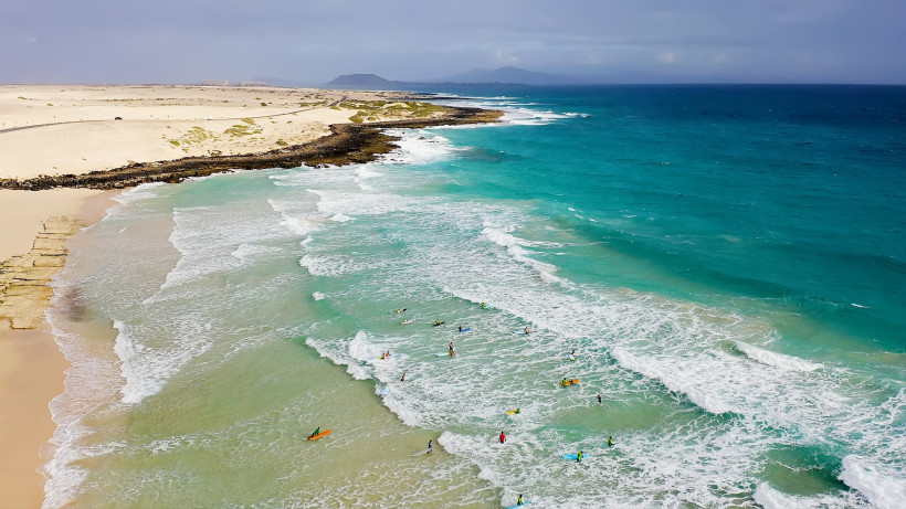 Küstenabschnitt bei Corralejo mit Dünenstrand, türkisblauem Meer und Brandung; mehrere Surfer im Wasser