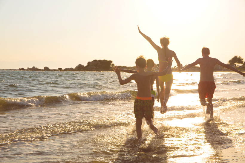 Vierköpfige Familie läuft lachend durch das flache Wasser am Strand, im Gegenlicht der tief stehenden Sonne.