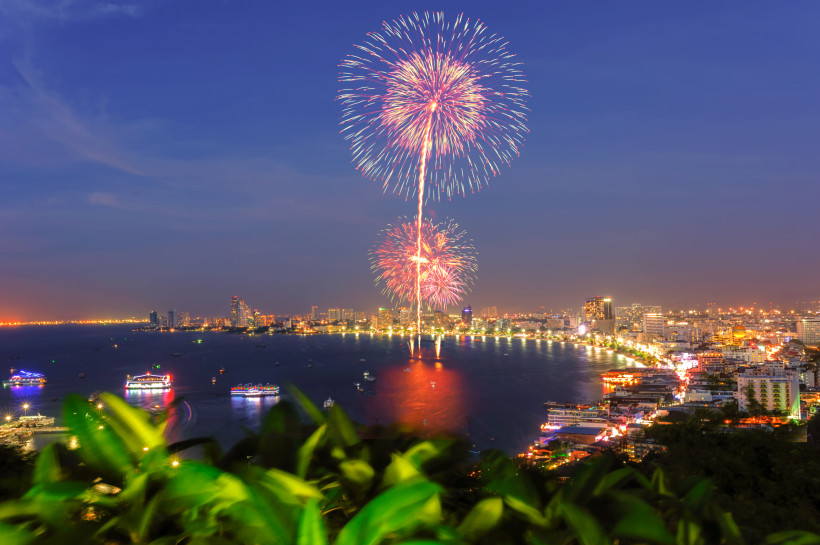 Thailand Pattaya Farbenfrohes Feuerwerk über der Bucht von Pattaya bei Nacht. Im Vordergrund grüne Pflanzen, im Hintergrund leuchtende Boote auf dem Wasser und die hell erleuchtete Skyline der Stadt.