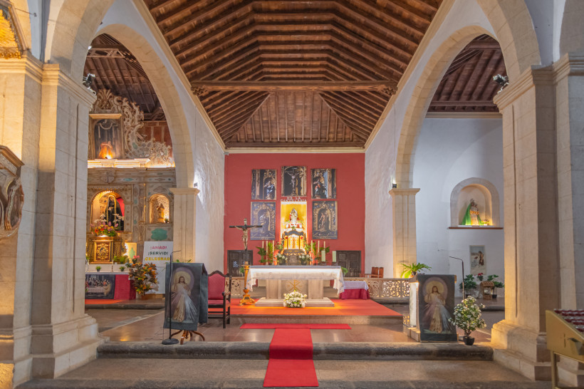Fuerteventura Blick in das Innere einer Kirche mit einem festlich geschmückten Altar, roten Teppichen, Heiligenfiguren und religiösen Gemälden. Das Gewölbe ist aus Holz, flankiert von steinernen Bögen.