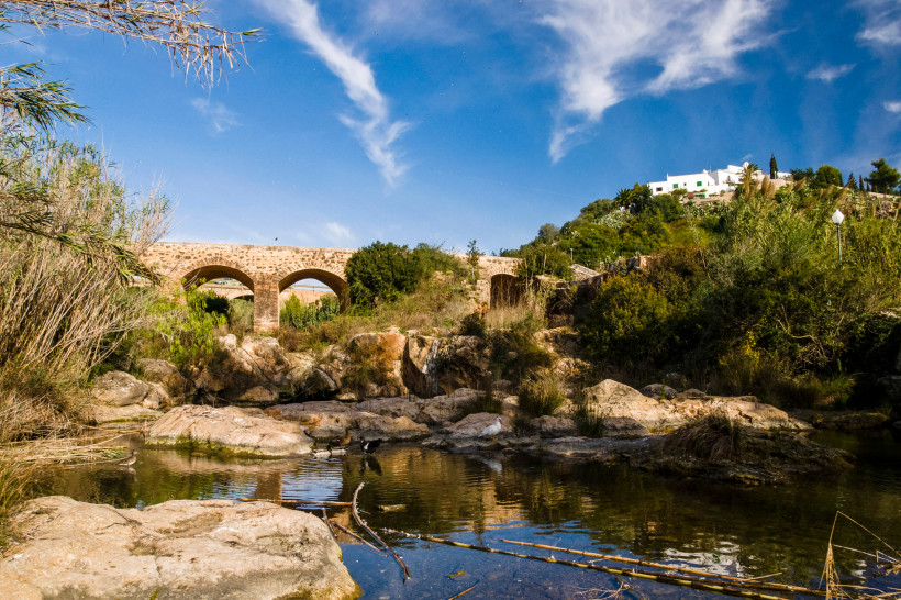 Santa Eulària des Riu Historische Steinbrücke mit mehreren Bögen über einem flachen Flussbett, umgeben von Felsen, Sträuchern und Bäumen. Auf einem Hügel im Hintergrund steht ein weißes Haus vor blauem Himmel.