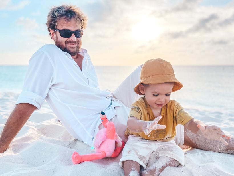 Ein Mann mit Sonnenbrille und weißem Hemd sitzt entspannt im weißen Sand am Strand von Aruba. Neben ihm spielt ein Kleinkind mit Sand, trägt einen beigefarbenen Sonnenhut und ein gelbes Shirt mit Auto-Motiven. Zwischen ihnen liegt ein pinkes Stoff-Flaming