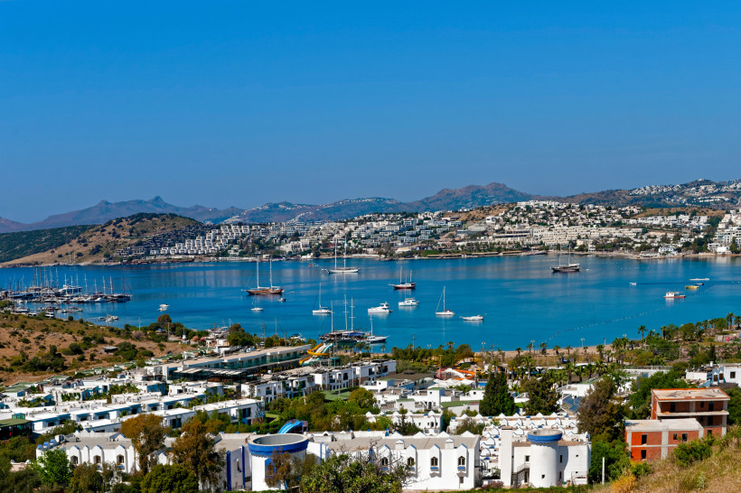 Panoramablick auf eine mediterrane Bucht mit Yachthafen, weißen Hotelanlagen und Segelbooten vor hügeliger Küstenlandschaft