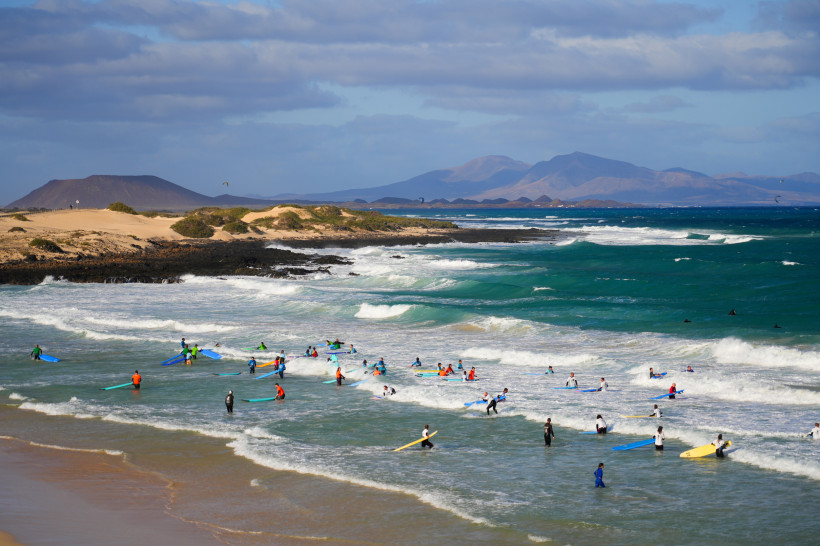 Strand bei Corralejo mit Surfern im Brandungswasser, Sanddünen und Bergen im Hintergrund
