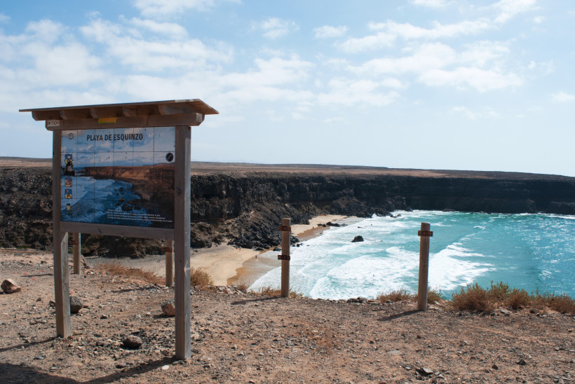 Informationstafel zur Playa de Esquinzo mit Blick auf eine felsige Bucht und Sandstrand am Meer