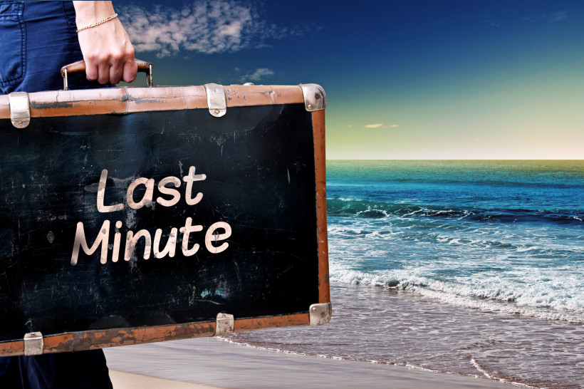 Person mit Koffer am Strand mit der Aufschrift „Last Minute“ vor Meer und Himmel – Symbolbild für Last-Minute-Urlaub und Spontanreisen
