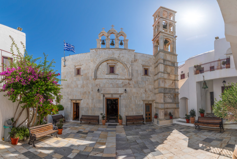 Heller Innenhof mit einer traditionellen griechischen Kirche aus hellem Stein. Über dem Eingang befinden sich drei Glocken unter kleinen Bögen. Rechts steht ein Glockenturm, im Hof blühen violette Bougainvillea-Blumen. Griechische Flagge weht im Hintergru