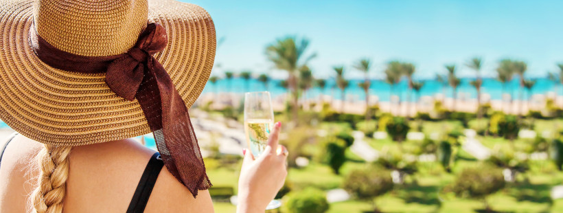 Ägypten Frau mit langem blondem Zopf und breitem Sonnenhut blickt mit einem Glas Sekt in der Hand auf ein tropisches Resort mit Palmen und Meer im Hintergrund.