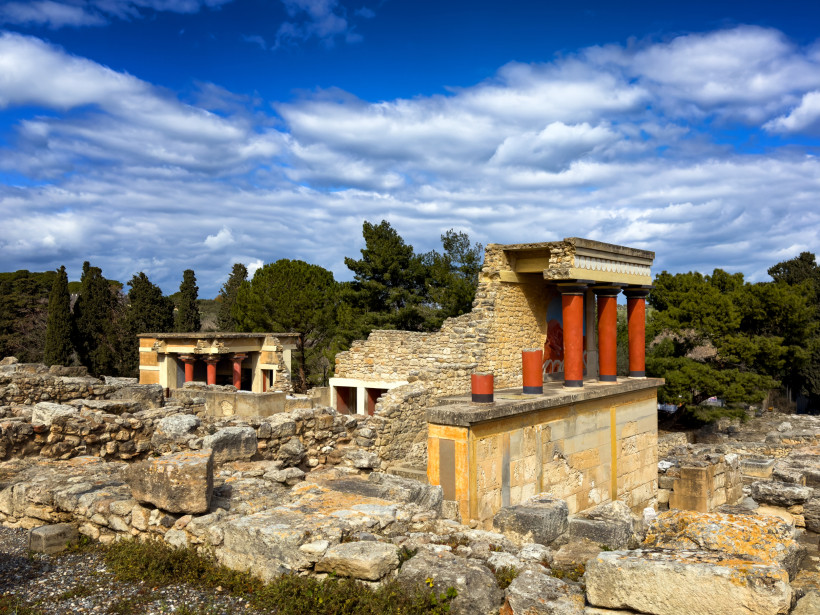Palastanlage von Knossos mit rekonstruierten roten Säulen und Steinruinen Rekonstruktion mit roten Säulen in der Palastanlage von Knossos, umgeben von Ruinen und Bäumen unter bewölktem Himmel