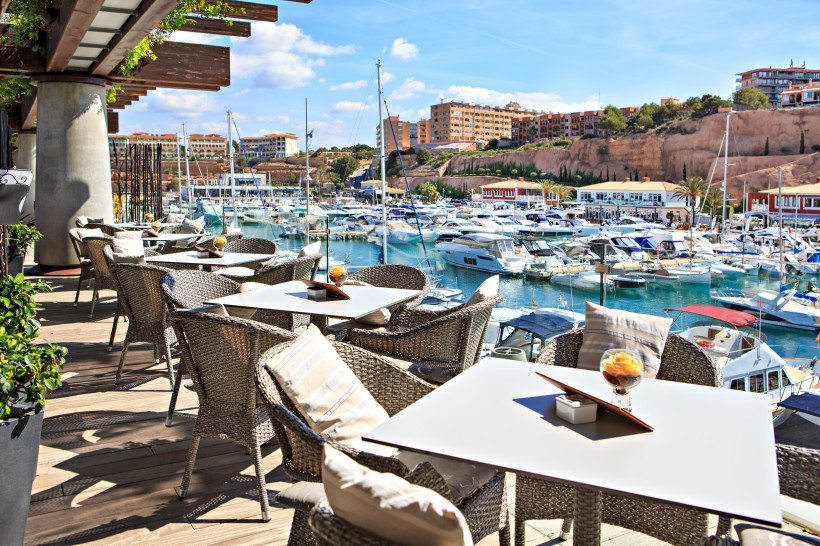 Terrasse mit Tischen und Stühlen am Yachthafen Port Adriano, im Hintergrund zahlreiche Boote und Gebäude am Hang