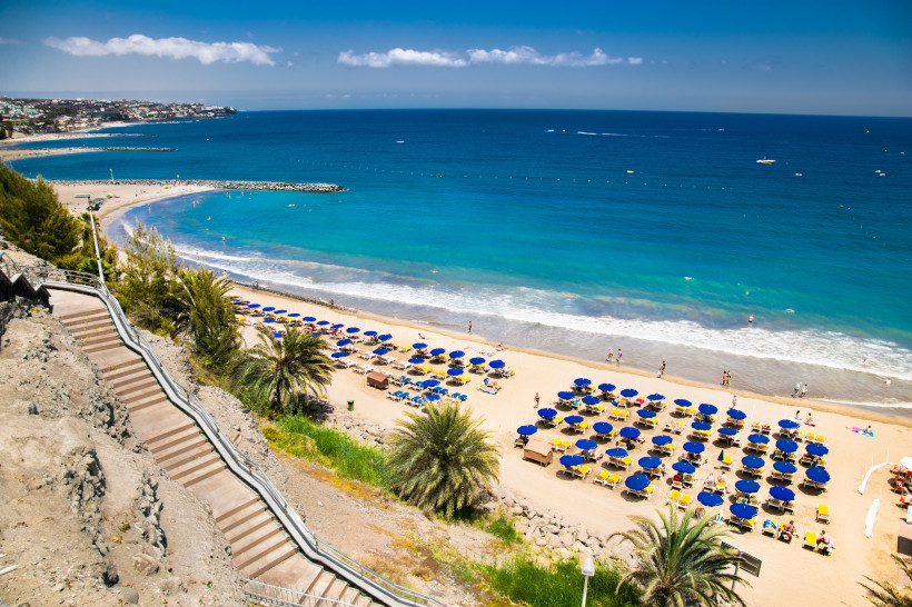 San Agustín Strand auf Gran Canaria – Goldgelber Sandstrand mit türkisblauem Meer Panoramablick auf den Strand von San Agustín im Süden Gran Canarias mit goldgelbem Sand, türkisblauem Meer und Sonnenschirmen. Eine breite Treppe führt hinunter zur Küste, während Urlauber im Wasser und am Strand entspannen.