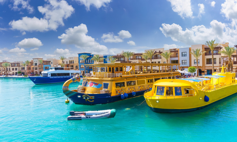 Ägypten - Marsa Alam Bunte Ausflugsboote im türkisfarbenen Wasser eines Yachthafens. Im Vordergrund ein gelbes Glasbodenboot und ein ägyptisch gestaltetes Ausflugsboot. Dahinter Ferienhäuser mit Palmen und blauer Himmel.