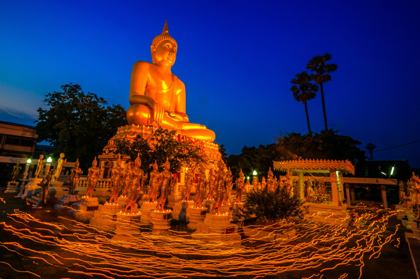 Thailand Das Bild zeigt eine eindrucksvoll beleuchtete, goldene Buddha-Statue bei Nacht. Sie ist von zahlreichen kleineren stehenden Buddhafiguren umgeben. Im Vordergrund verlaufen leuchtende Linien über den Boden – entstanden durch lange Belichtungszeit, während