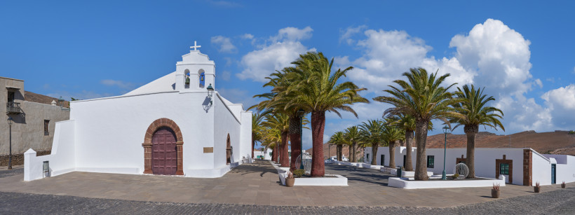 Lanzarote Weiße Kirche mit Glockengiebel und brauner Holztür auf einem sonnigen Platz mit Palmen. Umgeben von weiß getünchten Häusern unter blauem Himmel mit vereinzelten Wolken