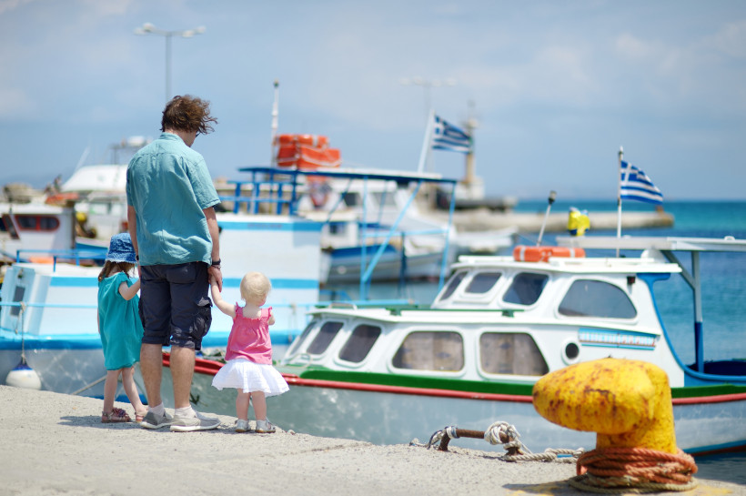 Ein Mann steht mit zwei kleinen Kindern an einem Hafen mit griechischen Fischerbooten. Die Gruppe ist von hinten zu sehen. Die Kinder halten seine Hände – das eine trägt ein blaues Kleid und einen Sonnenhut, das andere ein pinkes Top mit weißem Rock. Im H
