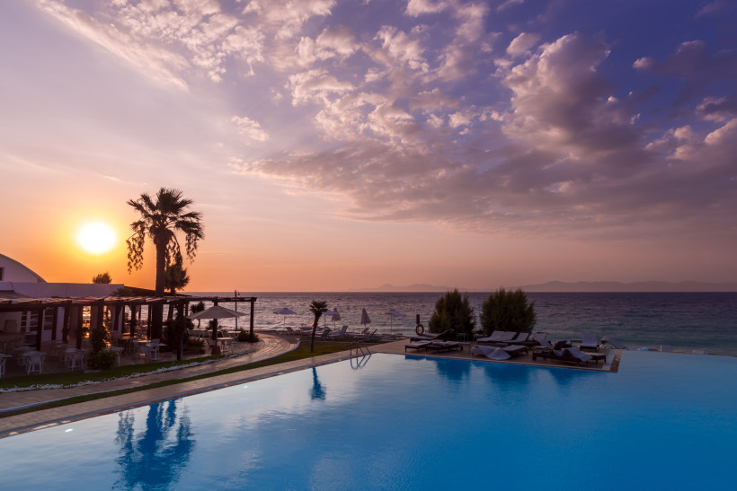 Infinity-Pool eines Strandhotels bei Sonnenuntergang mit Palmen, Liegen und Blick auf das Meer in Griechenland