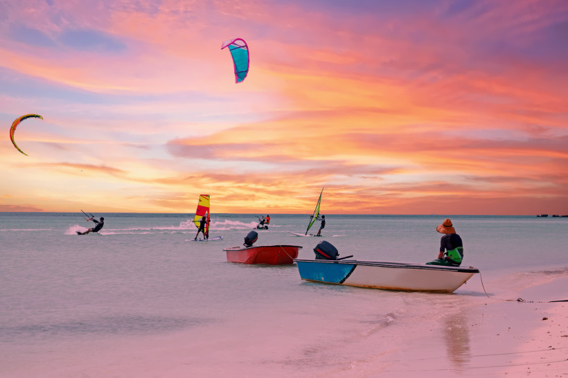 Mehrere Personen beim Kitesurfen und Windsurfen auf ruhigem Wasser vor Aruba bei Sonnenuntergang. Der Himmel leuchtet in warmen Farben von Orange bis Lila. Drei kleine Boote liegen am Ufer, eine Person sitzt auf einem Boot und beobachtet das Geschehen.