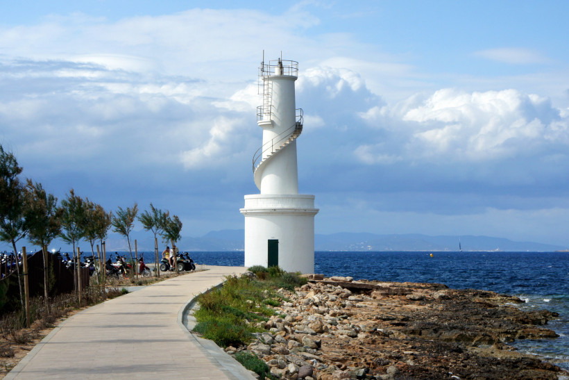 Formentera Weißer Leuchtturm mit spiralförmiger Außentreppe an einem Küstenweg, umgeben von Felsen, Büschen und dem Meer. Der Himmel ist leicht bewölkt, im Hintergrund sind Berge zu sehen.