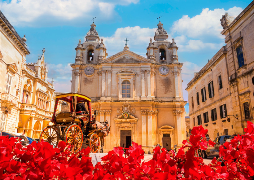 Mdina auf Malta – Barocke Kathedrale und Pferdekutsche in der stillen Stadt Barocke Kathedrale St. Paul in Mdina auf Malta mit Pferdekutsche im Vordergrund und roten Blumen unter blauem Himmel