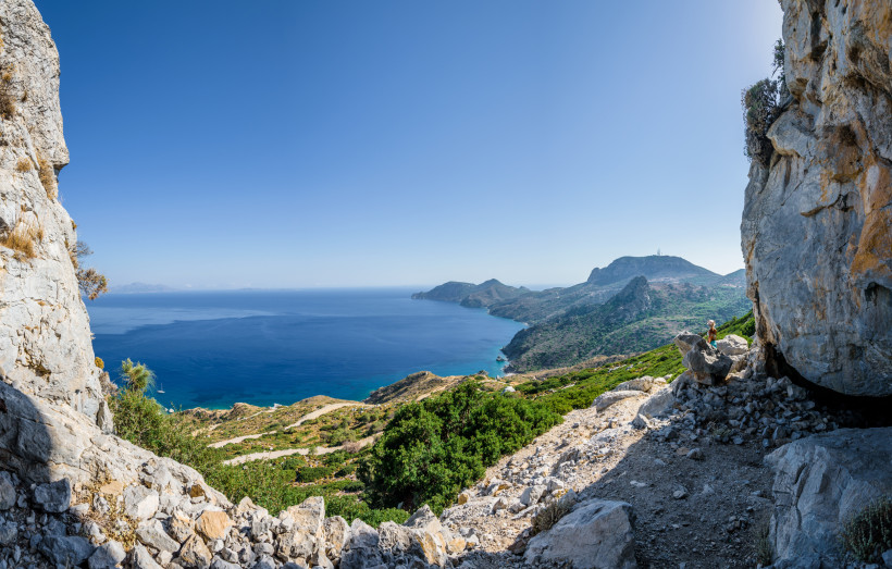Weitläufiger Panoramablick von einem felsigen Aussichtspunkt über eine grüne, bergige Küste. Im Vordergrund stehen große Felsen und eine einzelne Person mit Hut blickt auf die Landschaft. Unten ziehen sich Wege und Vegetation entlang der Berghänge. Das ti