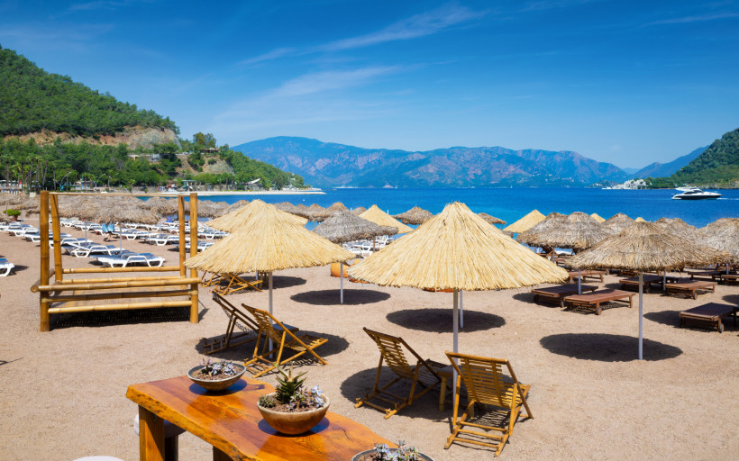 Ruhiger Strand in Icmeler mit Strohschirmen und Sonnenliegen. Ruhiges blaues Ägäisches Meer, malerische Berge unter klarem Himmel.