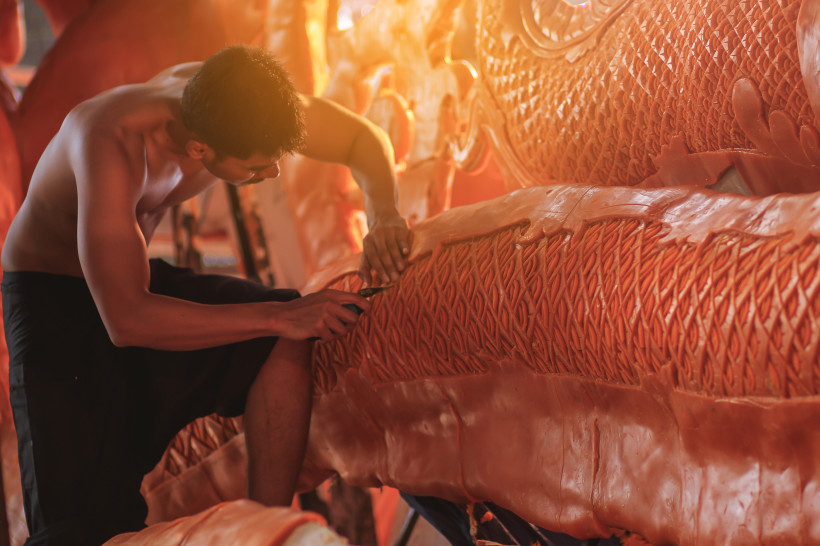 Ein junger Mann kniet konzentriert vor einer großen, kunstvoll geschnitzten Wachsskulptur in leuchtendem Gelb. Er trägt eine dunkle Hose, ist oberkörperfrei und arbeitet mit einem feinen Werkzeug an den Details